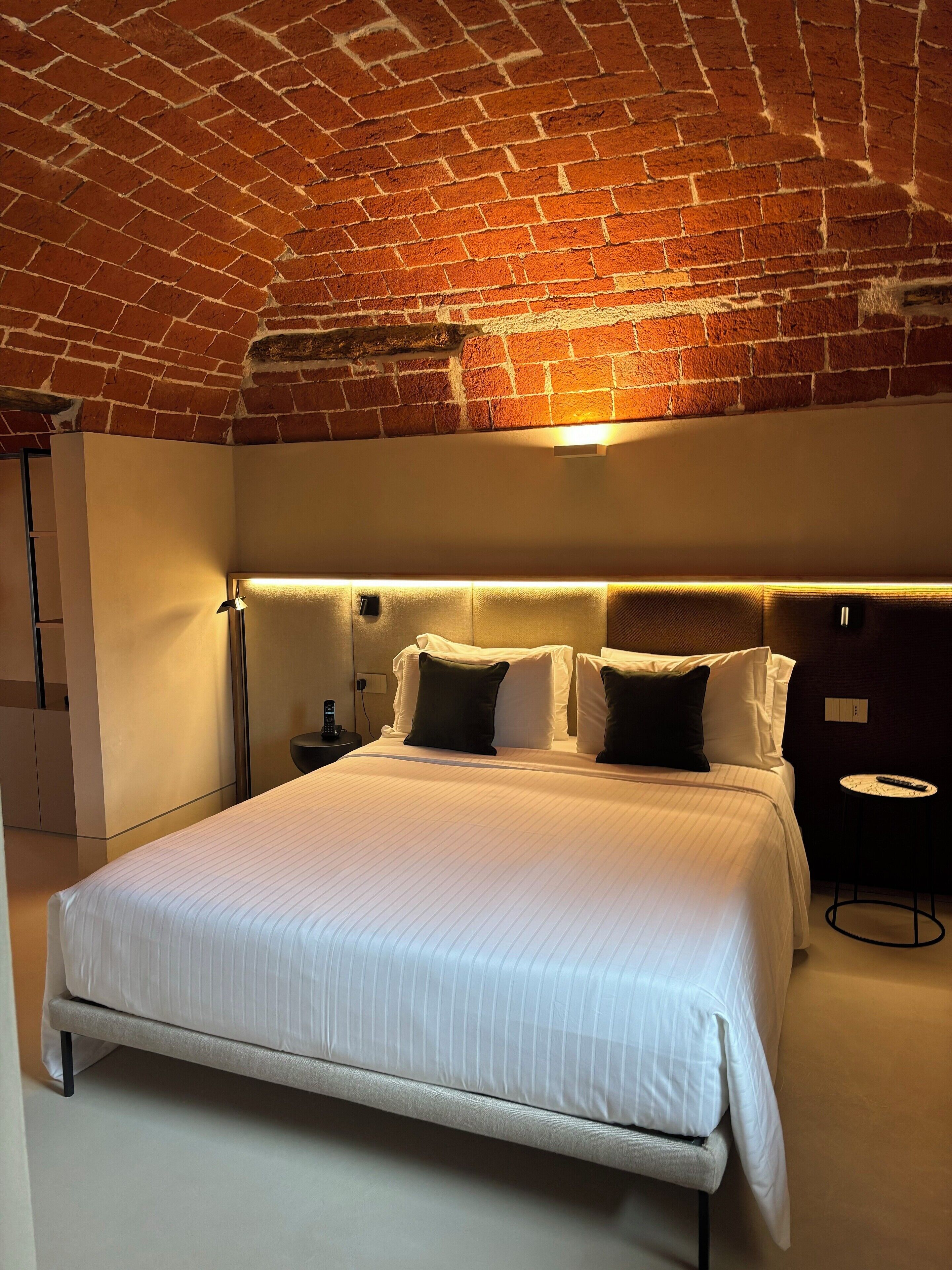 A modern hotel room with a brick ceiling, a large bed with white linens, a bedside table with a remote control, and a small round table.