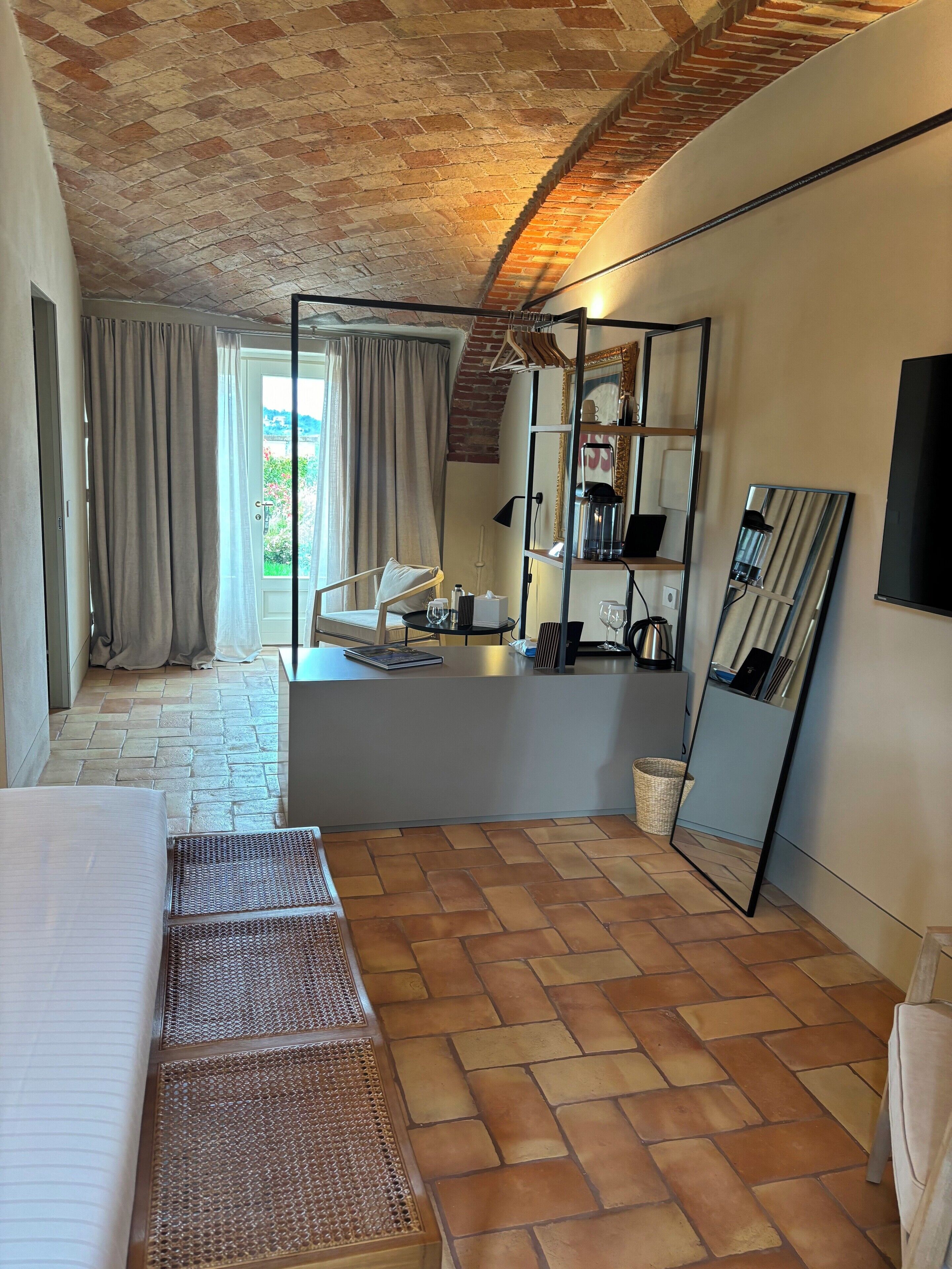 A room with a brick ceiling, a bed with a headboard, a small table with a lamp, a chair, and a large mirror.