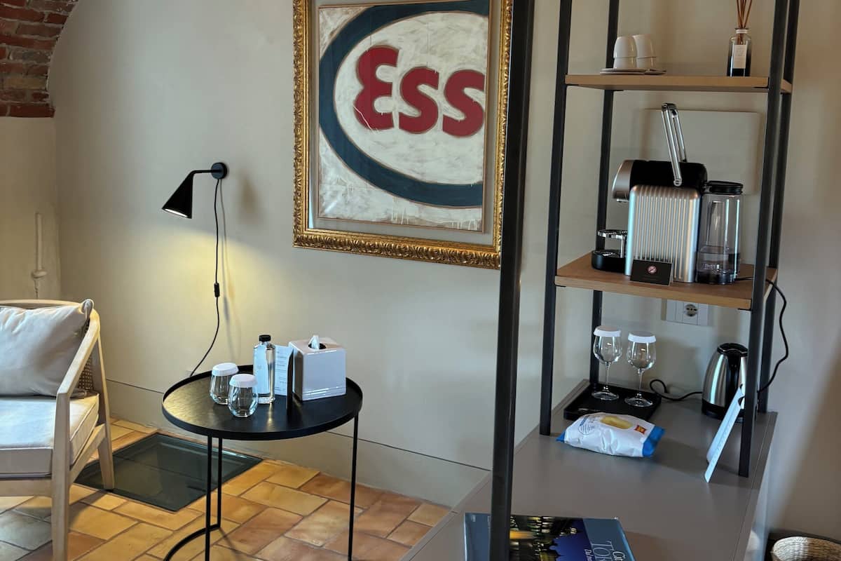 A cozy room with a brick wall, a framed 'ESS' sign, a round table with a lamp, a hanging rack, and a bookshelf with various items.