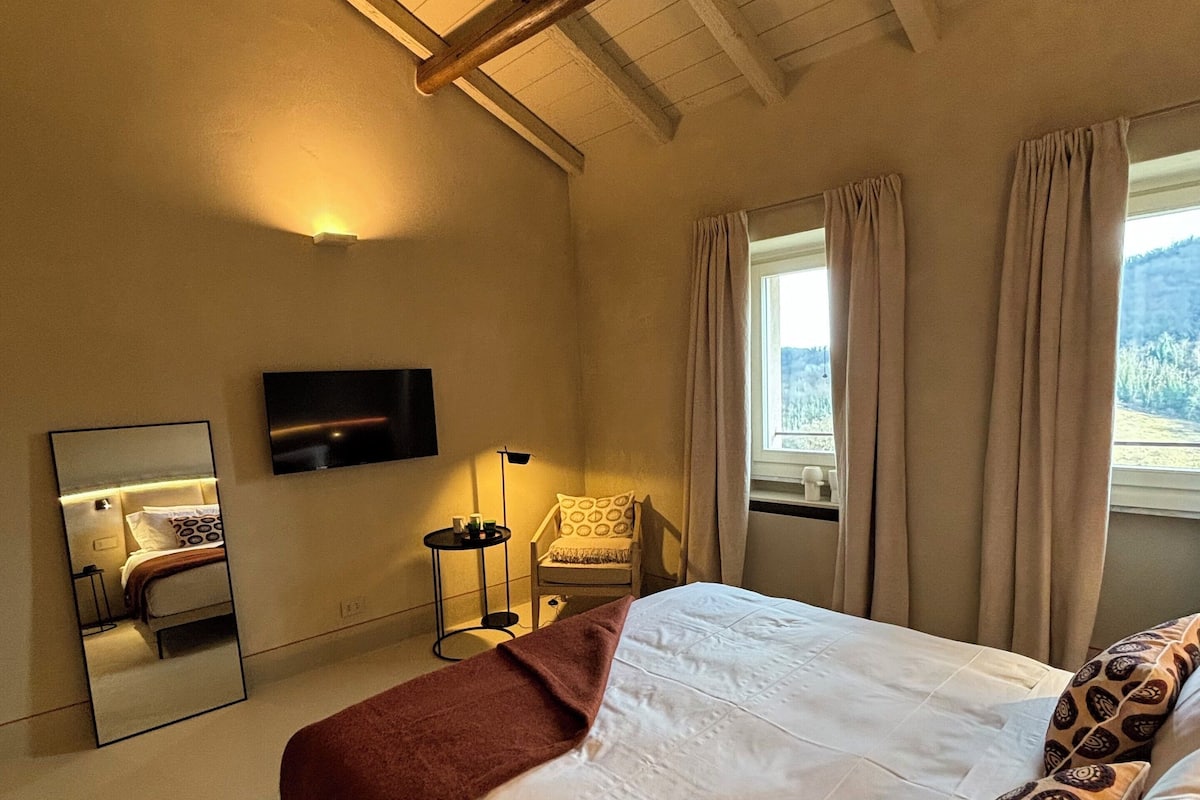 A bedroom with a large bed, a chair, a TV mounted on the wall, and a window with curtains.