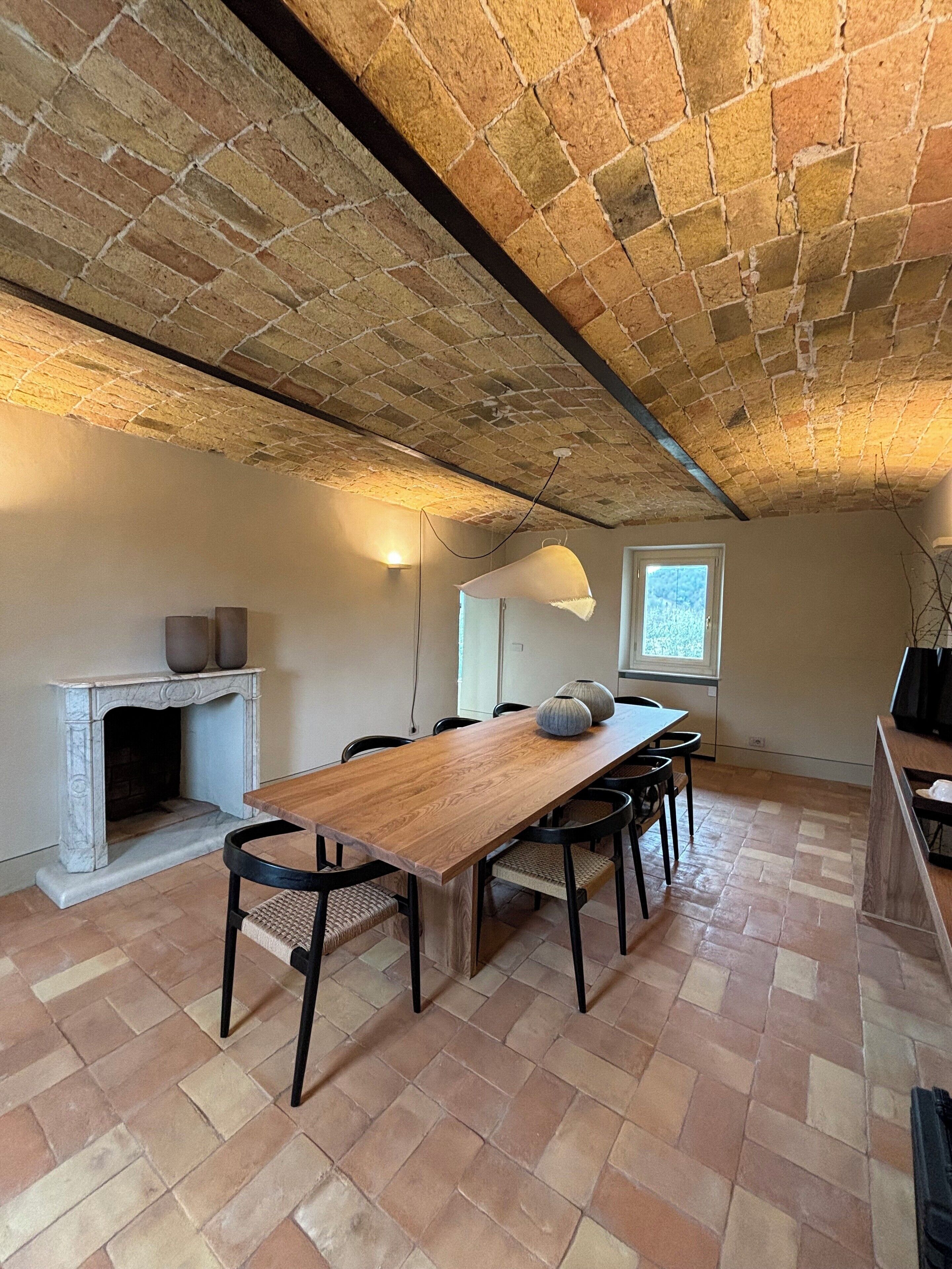 A dining area with a long wooden table, black chairs, a fireplace, and a brick ceiling.
