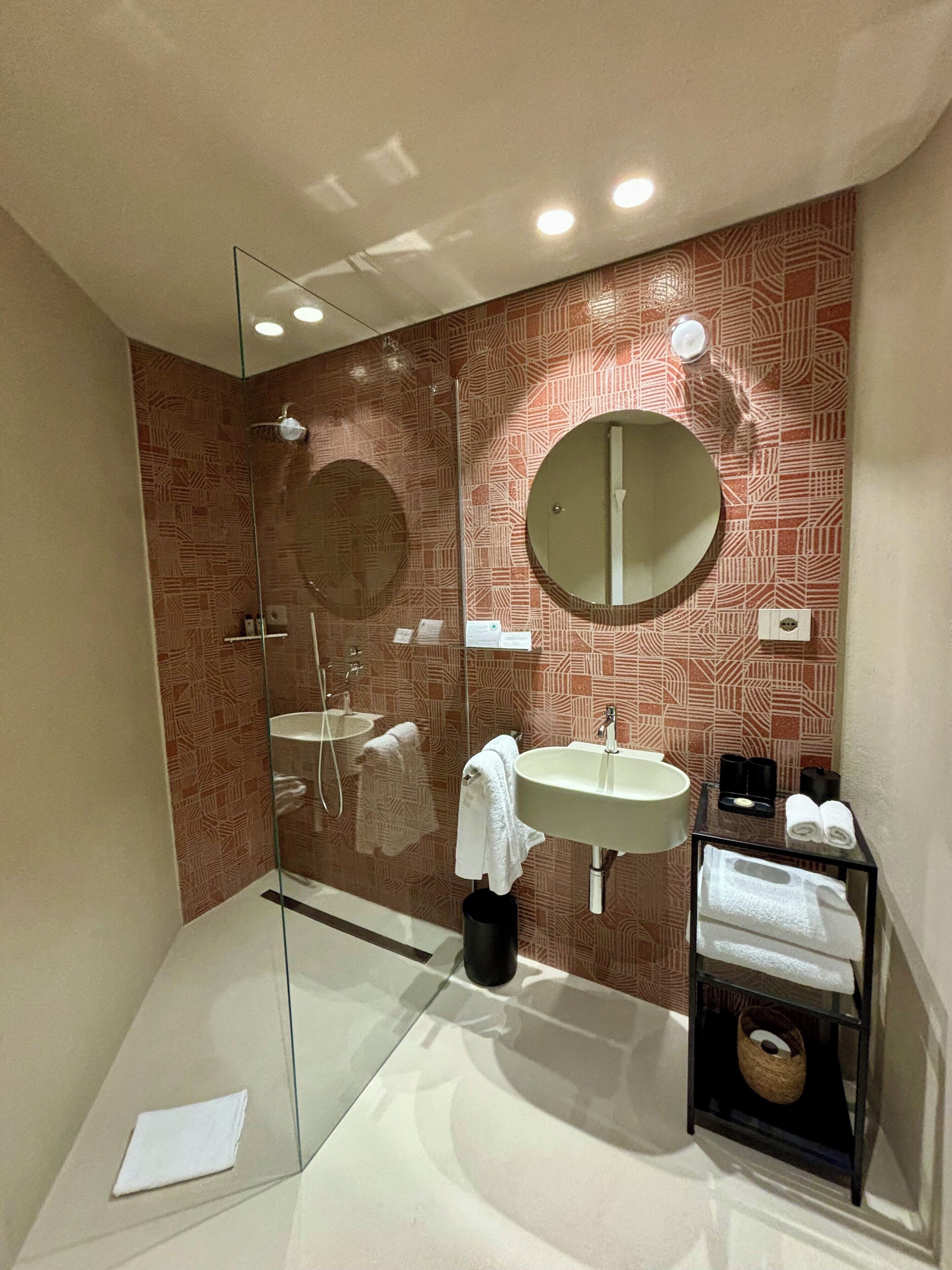 Un baño moderno con ducha de vidrio, un espejo redondo, un lavamanos y un perchero para toallas.