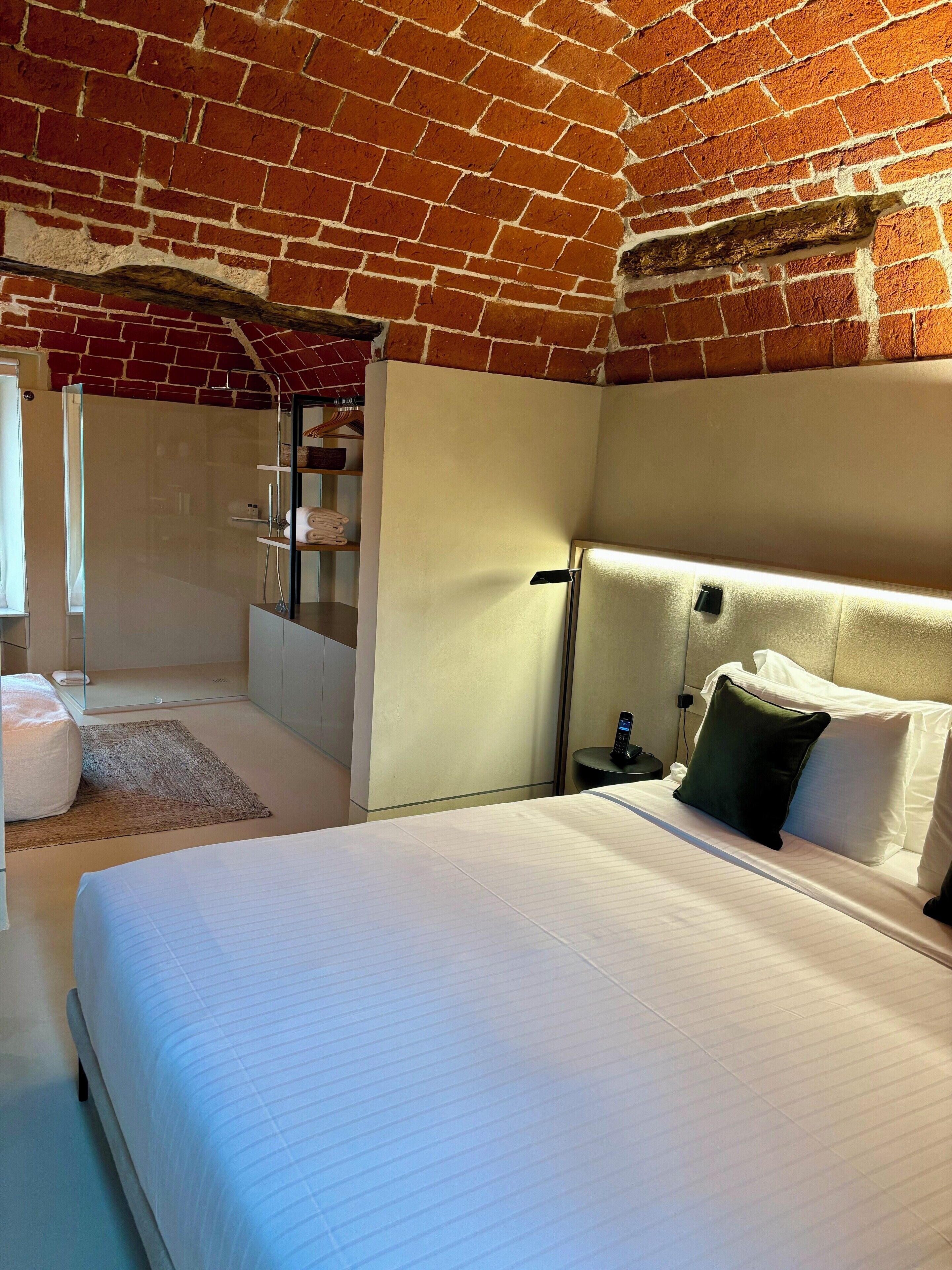 Una camera d'albergo moderna con soffitto in mattoni, un letto grande con lenzuola bianche, un comodino con una lampada e un altro piccolo comodino con un telefono.