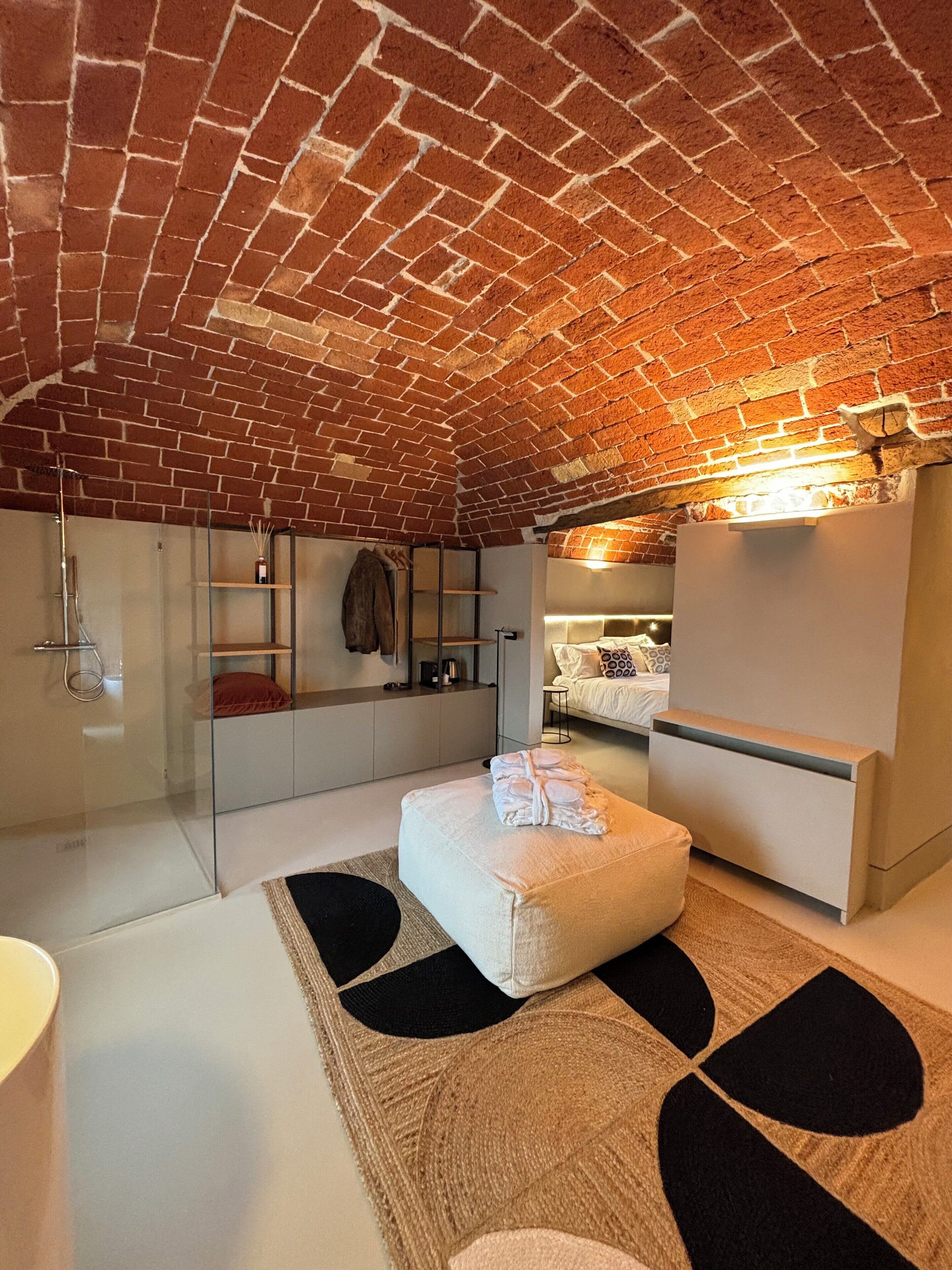 Un dormitorio tipo loft moderno con techo de ladrillo visto, cabina de ducha de vidrio, sofá blanco y cama con almohada estampada.
