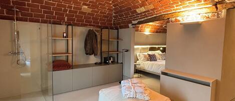 A modern loft bedroom with exposed brick ceiling, a glass shower enclosure, a white sofa, and a bed with a patterned pillow.