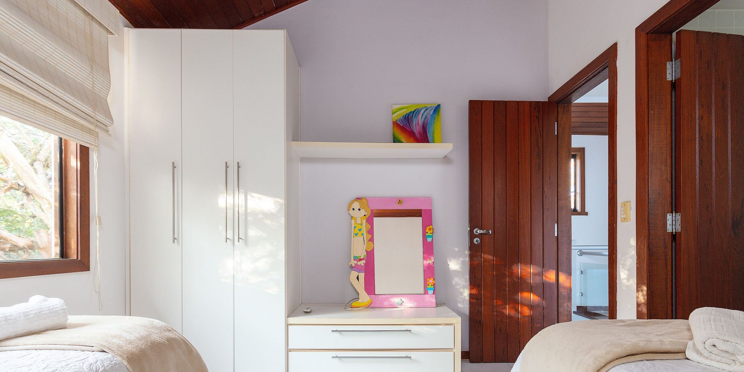 Une chambre dotée d’une grande armoire, d’une coiffeuse avec un miroir, d’une étagère ornée d’un tableau coloré et d’une porte donnant sur une autre pièce.