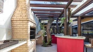 A brick wall, a grill, a tree, a red counter, and a wicker chair. - Bz55 Casa 200m da praia com 4 suítes (Buzios)