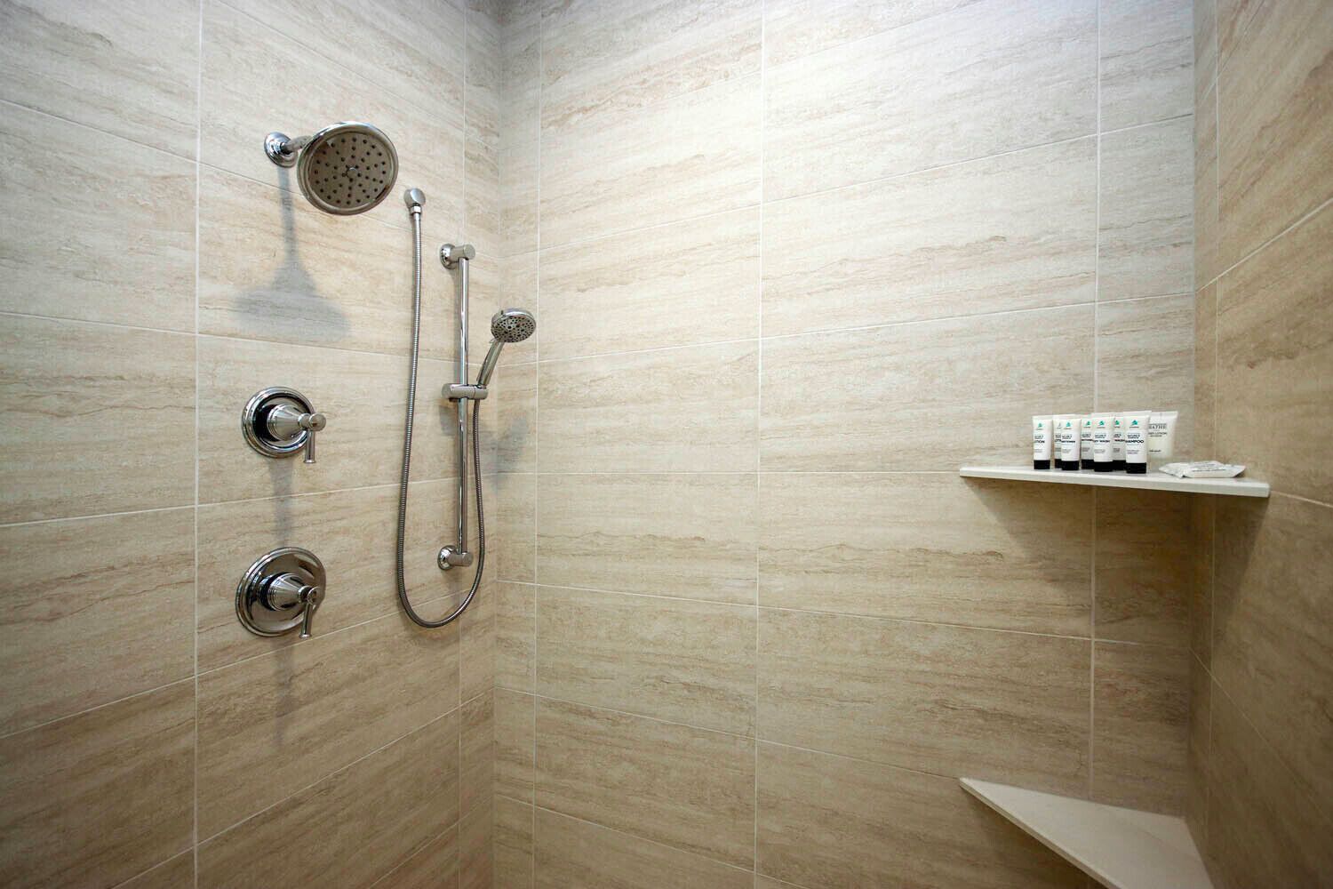 Un espace douche équipé d’un pommeau de douche à main, d’un pommeau de douche fixé au mur et d’un autre pommeau de douche à main avec un tuyau flexible. Une étagère contient des articles de toilette.