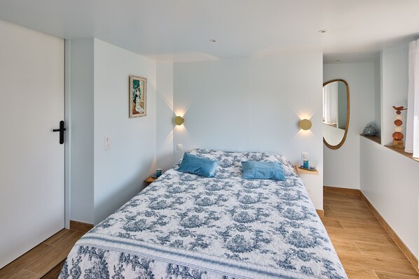 Camera da letto con un letto, due cuscini, un comodino, una lampada a parete, uno specchio e una mensola con oggetti decorativi.