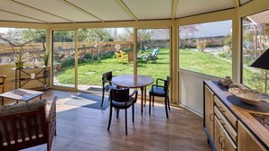 A sunlit room with a large sliding glass door leading to a garden. Inside, there is a wooden dining table with chairs, a small sofa, and a wooden cabinet with drawers. - Gîte between the Seine and the cliffs near GIVERNY and the Château de la Roche Guyon (Gommecourt)