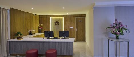A modern hotel lobby with a reception desk, two red stools, a small plant, and a decorative vase.