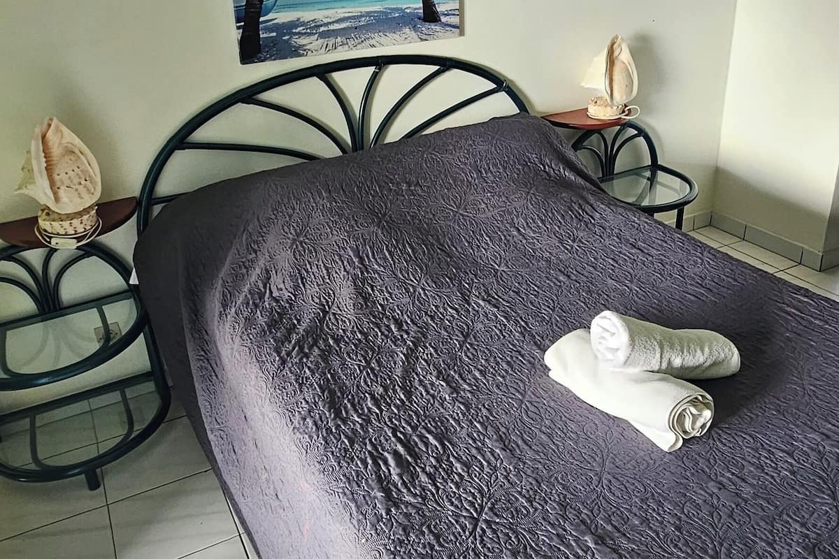 A neatly made bed with a dark textured bedspread, a small table with a lamp, and a wall with a tropical beach painting.
