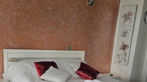Un lit bien fait, avec du linge de lit blanc et des coussins rouges, un plancher en bois et un mur à la finition texturée.