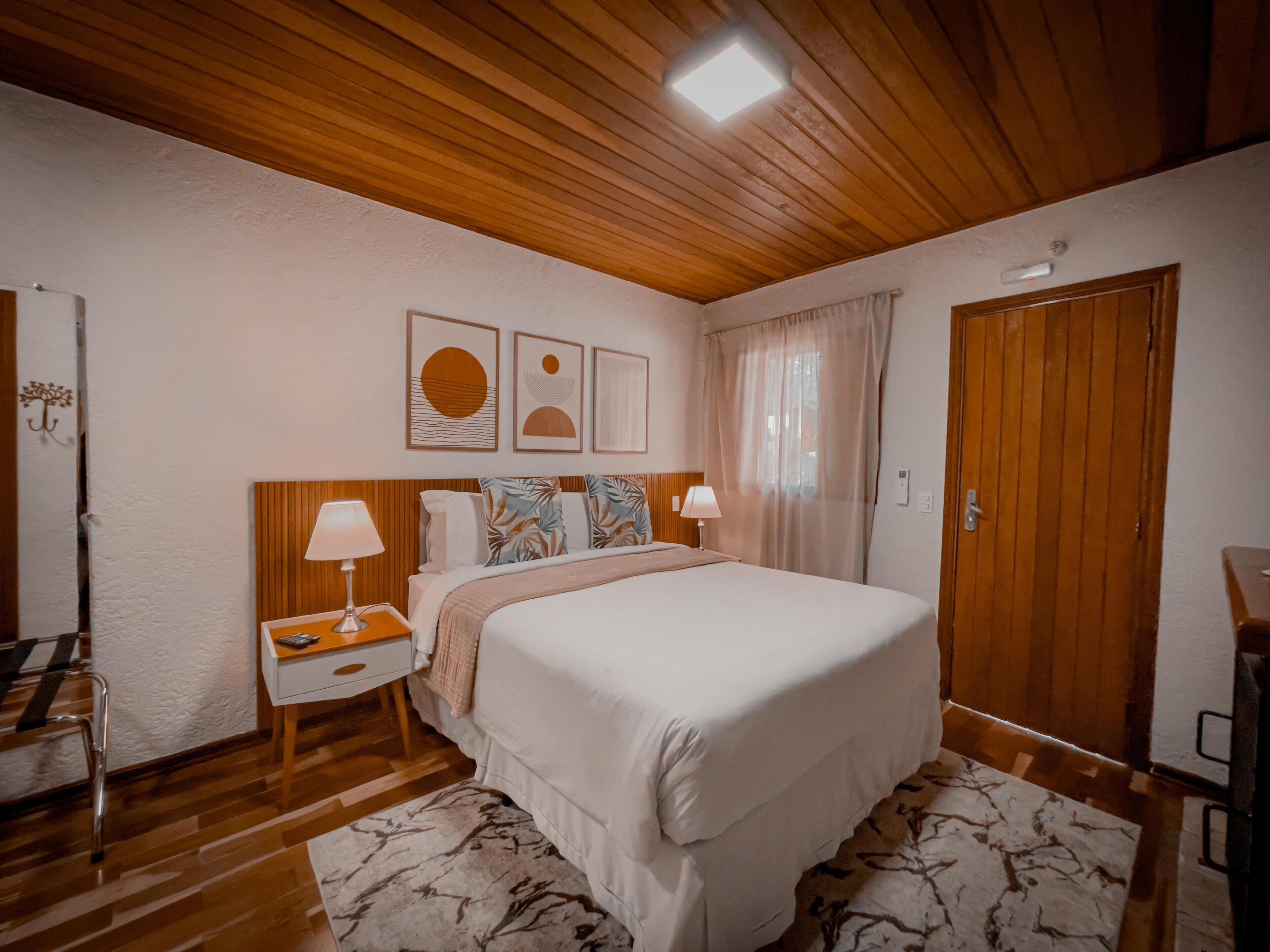Une chambre avec un plafond en bois, un lit avec une literie blanche, deux tables de chevet avec des lampes, une armoire en bois et un mur orné de trois œuvres d’art encadrées.