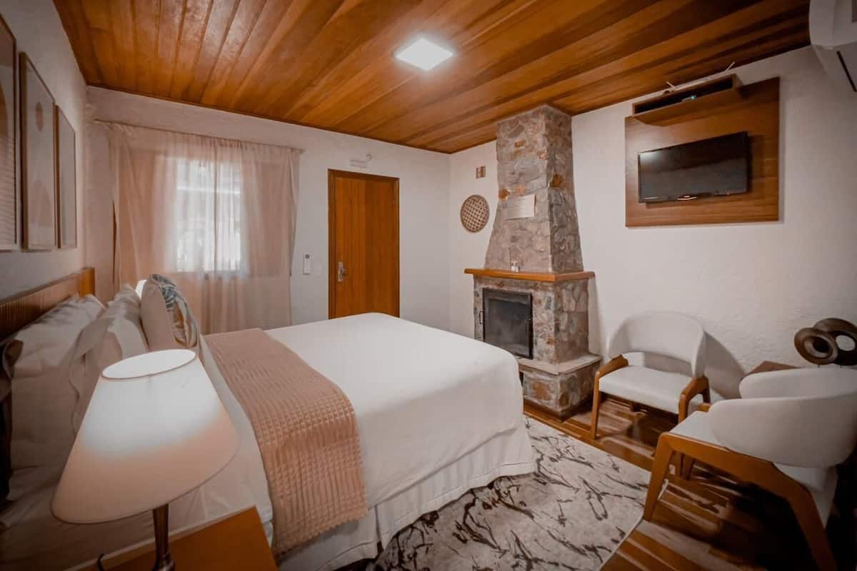 A bedroom with a large bed, a stone fireplace, a television mounted on the wall, and a wooden ceiling.