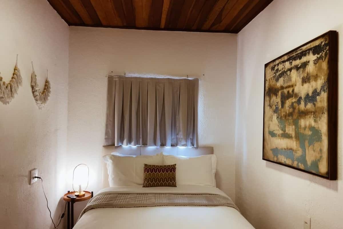 A bedroom with a bed, a bedside table with a lamp, a window with curtains, and a painting on the wall.