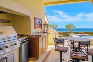 An outdoor kitchen with a Viking grill, a dining table with patterned chairs, and a view of the ocean.