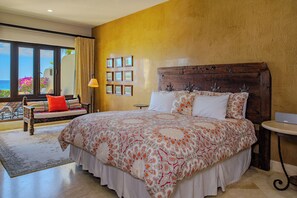 A bedroom with a large bed, a wooden headboard, a view of the ocean, a balcony with a chair, and a small table.
