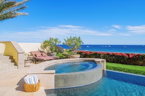 A curved infinity pool with a view of the ocean, a set of stairs, and outdoor seating with cushions.