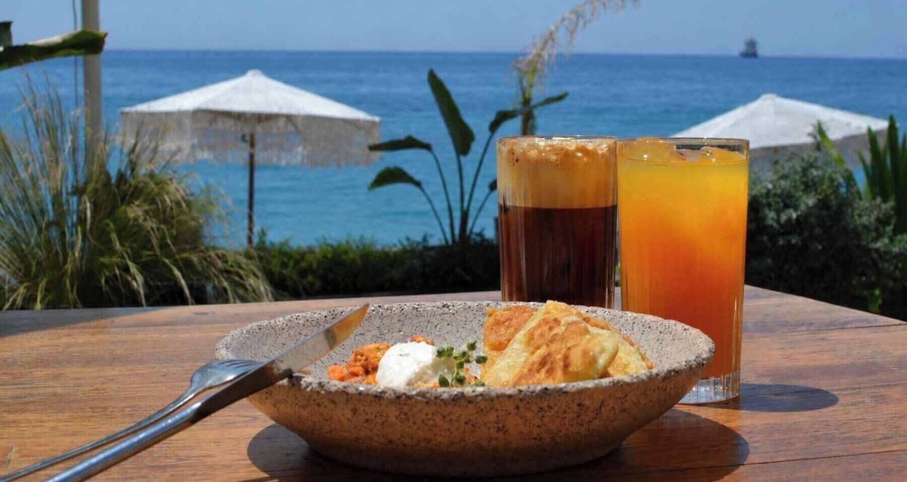 A table with a bowl of food and two drinks, a view of the sea, and umbrellas in the background.