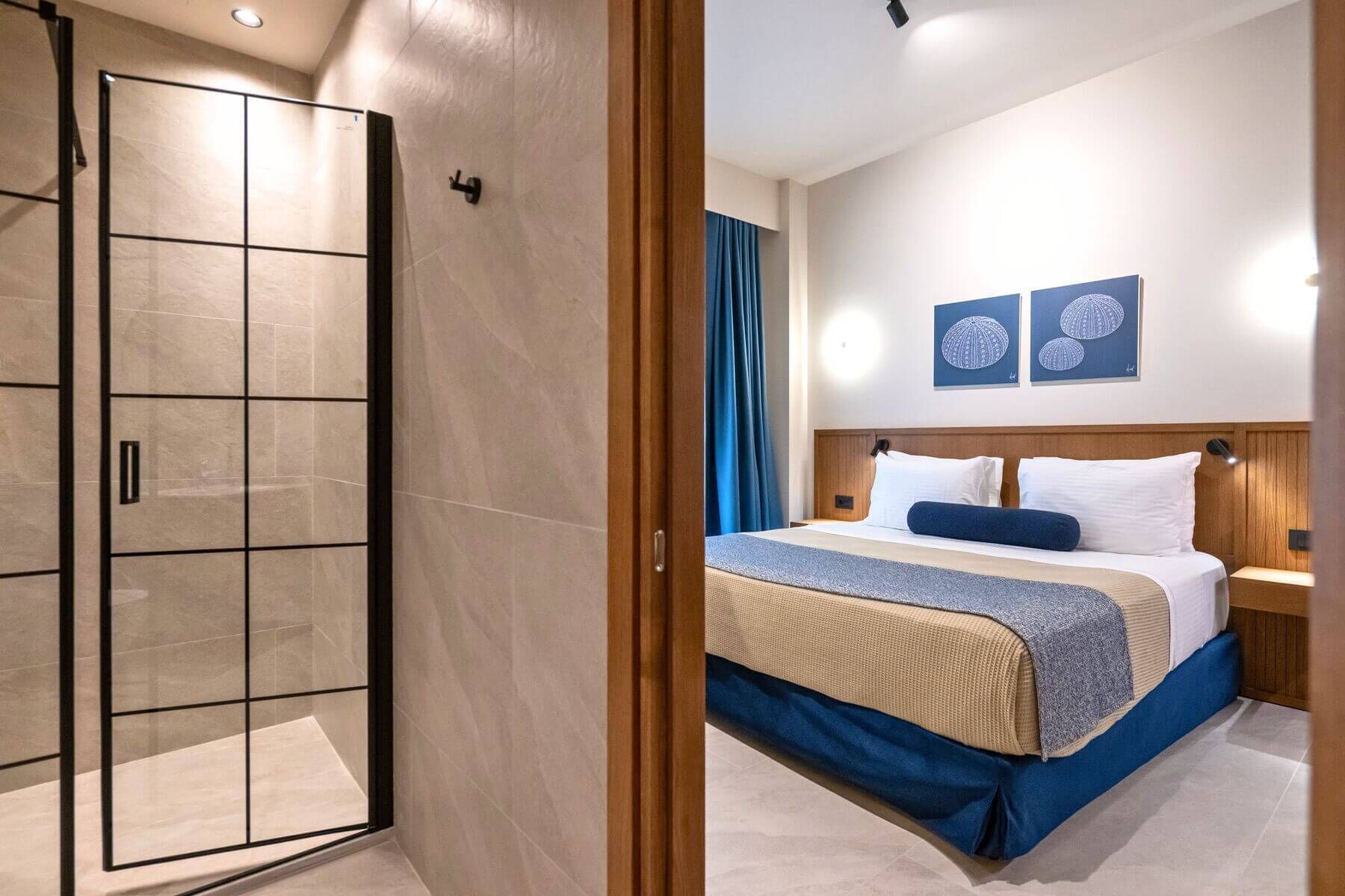 Een hotelkamer met een douchecabine van glas, een bed met houten hoofdbord en twee wandlampen.