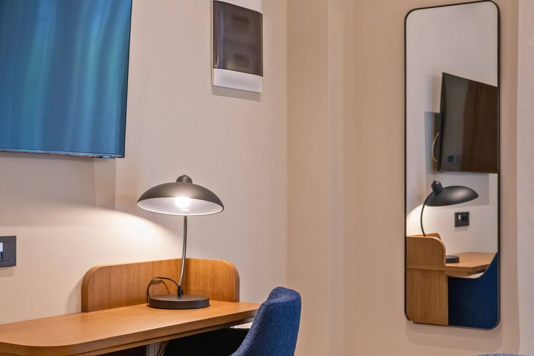A hotel room with a wooden desk, a blue chair, a desk lamp, a wall-mounted TV, and a mirror reflecting the room.