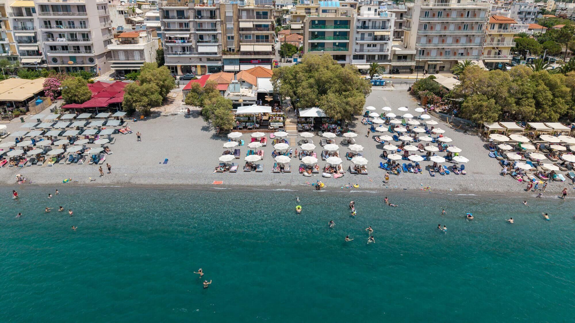 An aerial view of a beach with numerous umbrellas and sun loungers, a clear blue sea, and a densely packed urban backdrop.