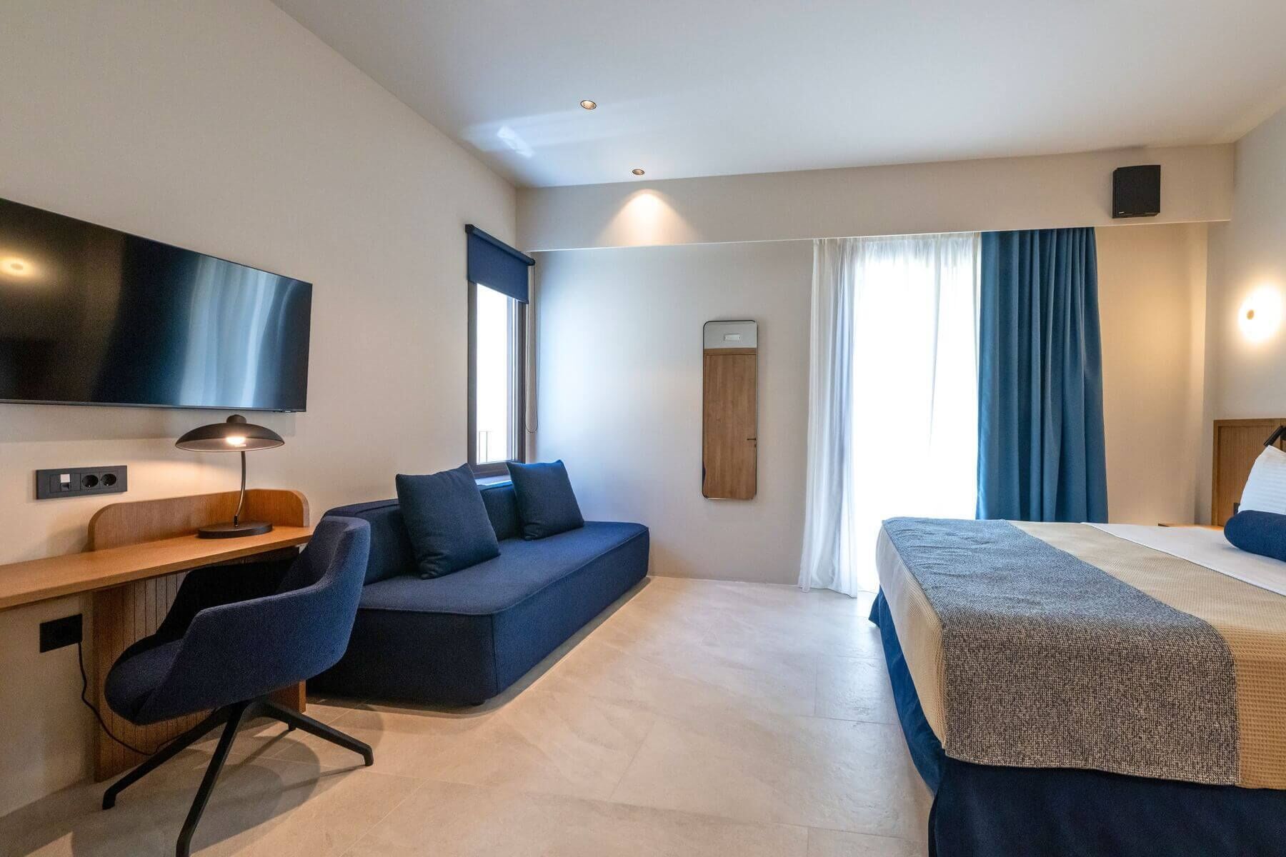A modern hotel room with a desk, a sofa, a bed, and a TV.