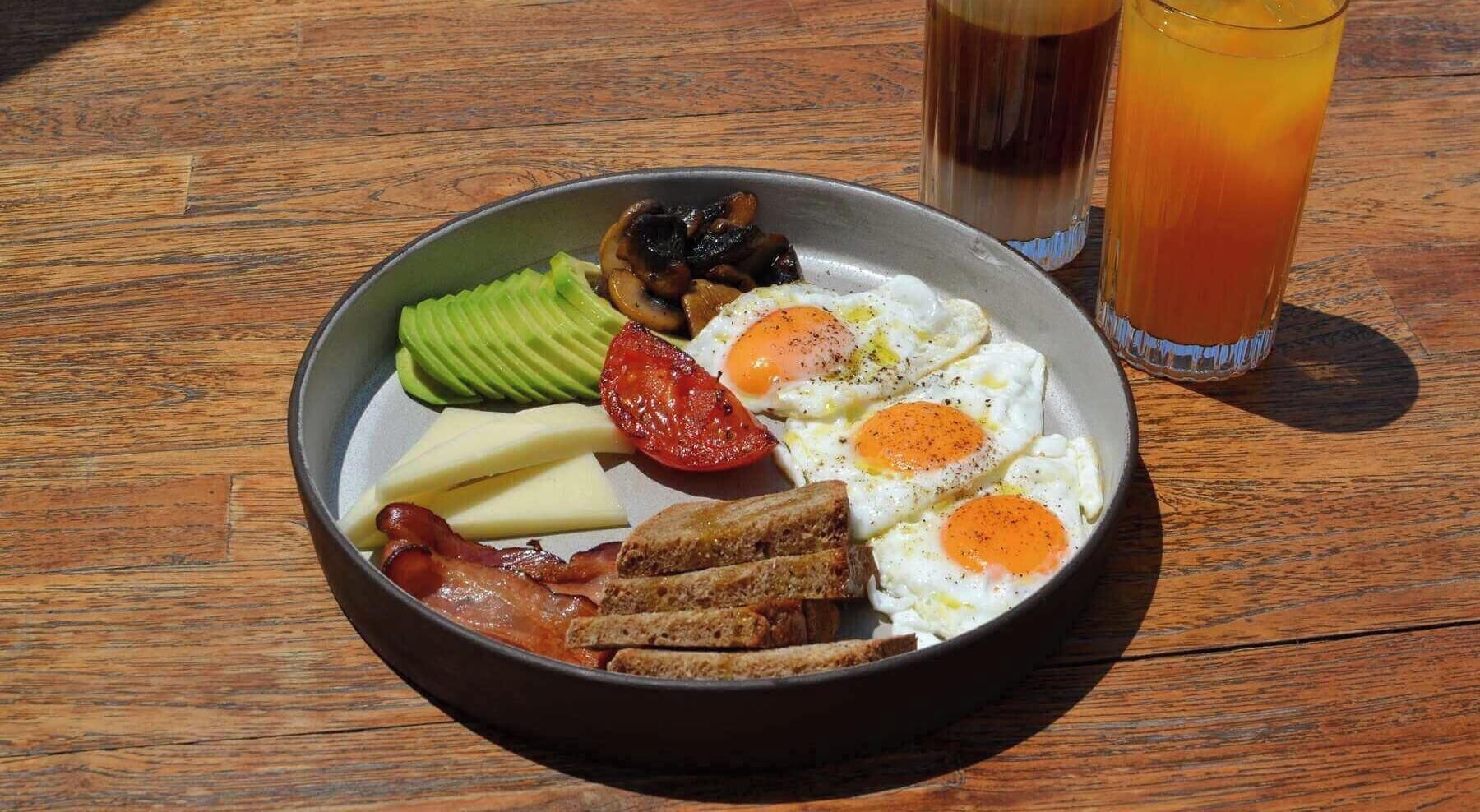A breakfast plate with eggs, bacon, avocado, tomato, cheese, and toast on a wooden table.