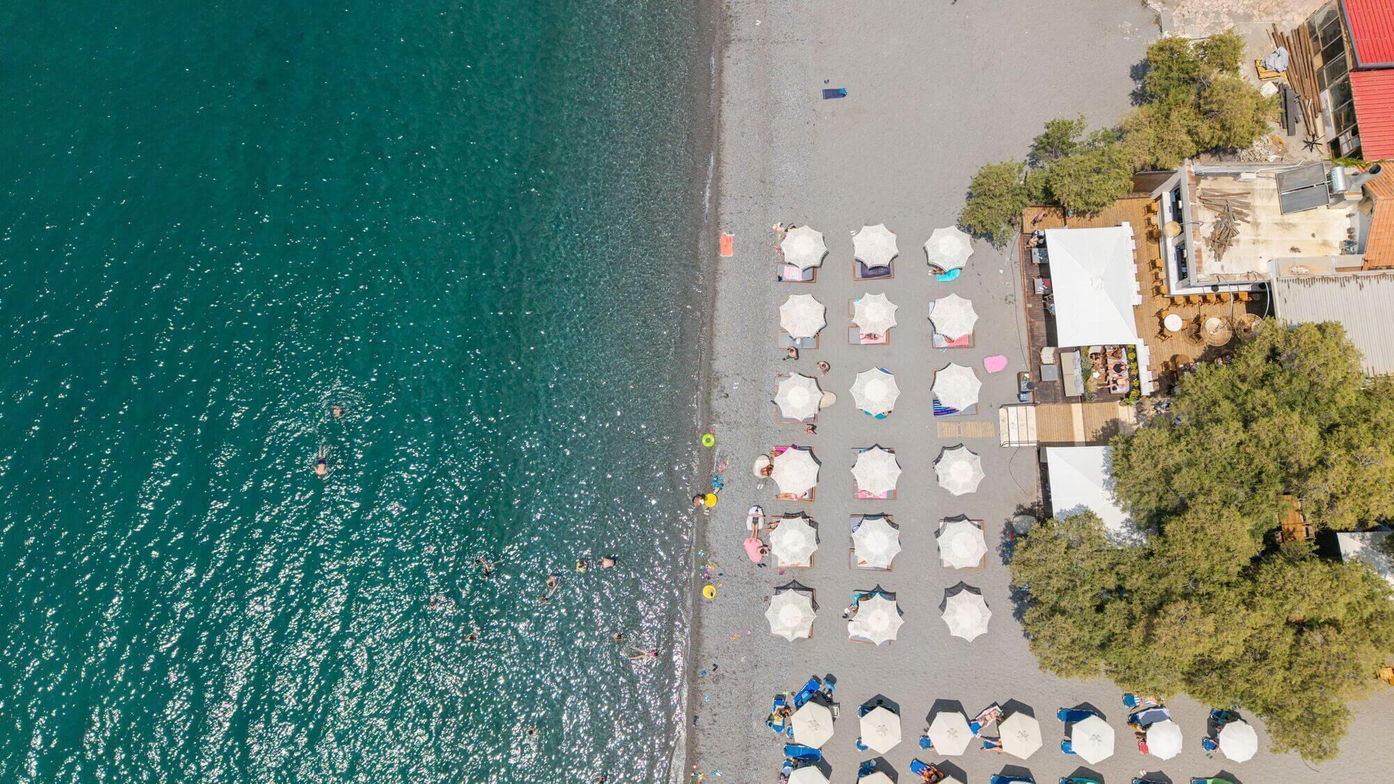 An aerial view of a beach with umbrellas and lounge chairs, a building with a red roof, and trees.