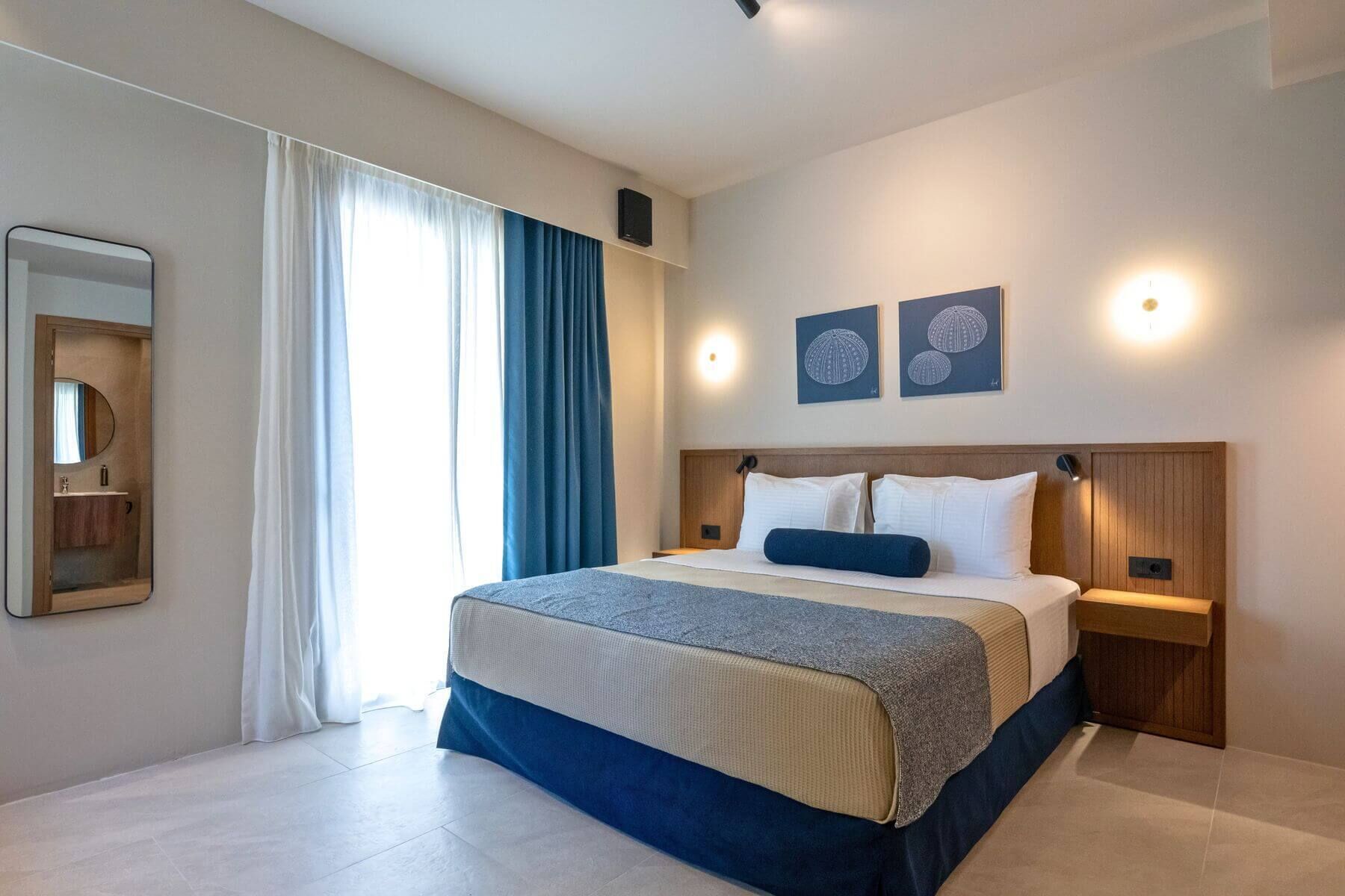 Quarto de hotel moderno com cama grande, mesa de cabeceira, espelho e janela com cortinas.