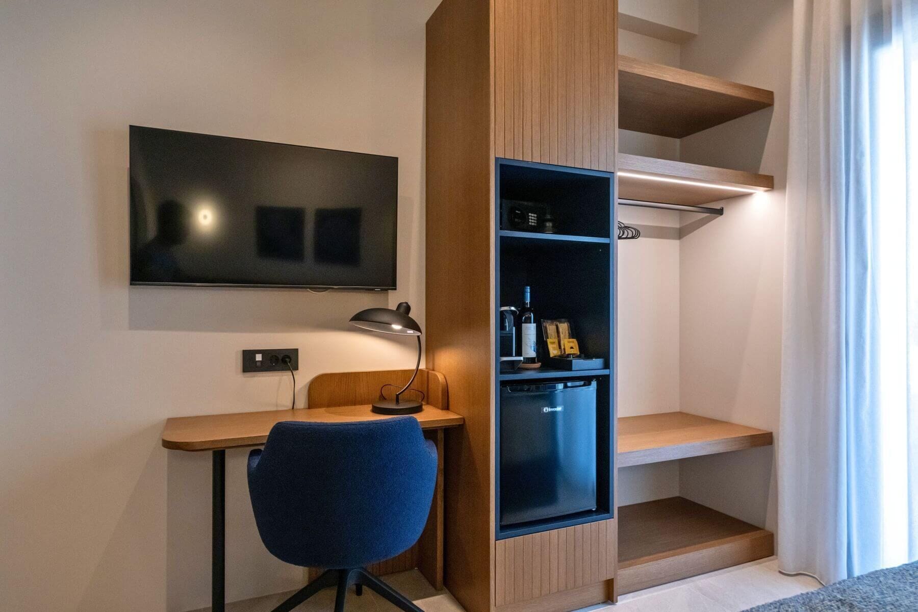 A modern hotel room with a flat-screen TV mounted on the wall, a desk with a chair, a built-in minibar, and a large window with curtains.