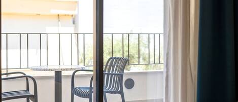 A balcony with a table and two chairs, a view of greenery outside, and a curtain partially covering the window.