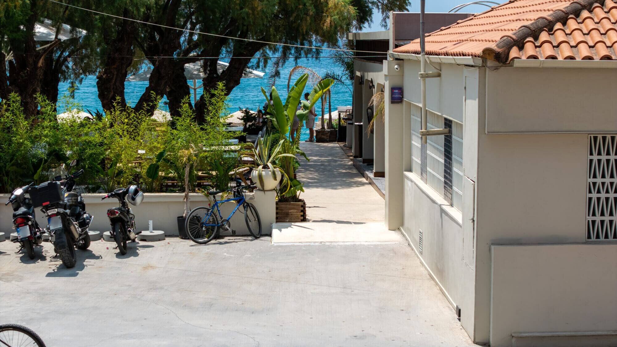 A narrow pathway leading to a pool area with a view of the sea. There are two motorcycles parked on the left and a bicycle on the right.