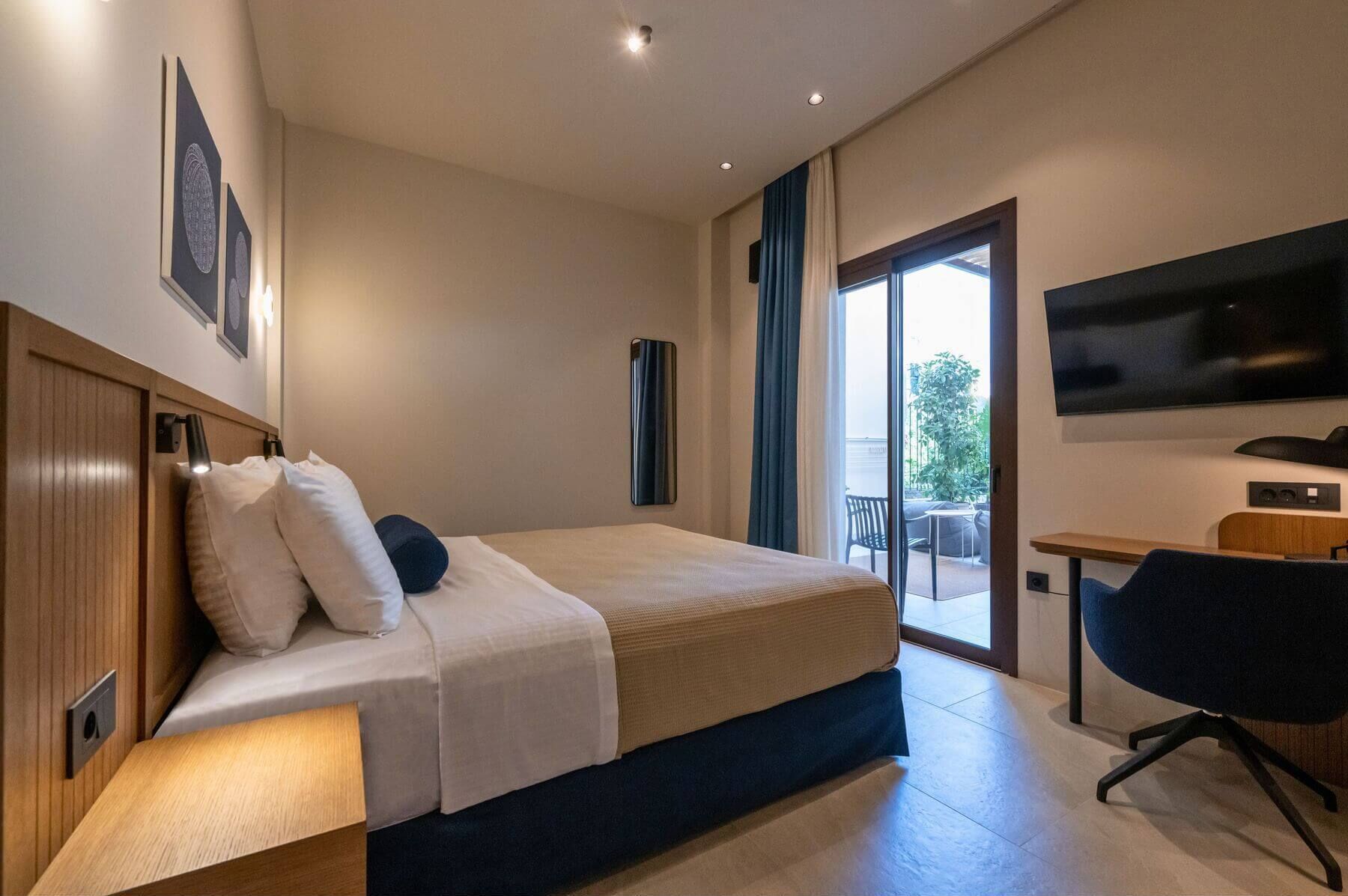 Une chambre d’hôtel moderne dotée d’un grand lit, d’un bureau, d’un téléviseur à écran plat et d’une porte-fenêtre coulissante donnant sur un espace extérieur.
