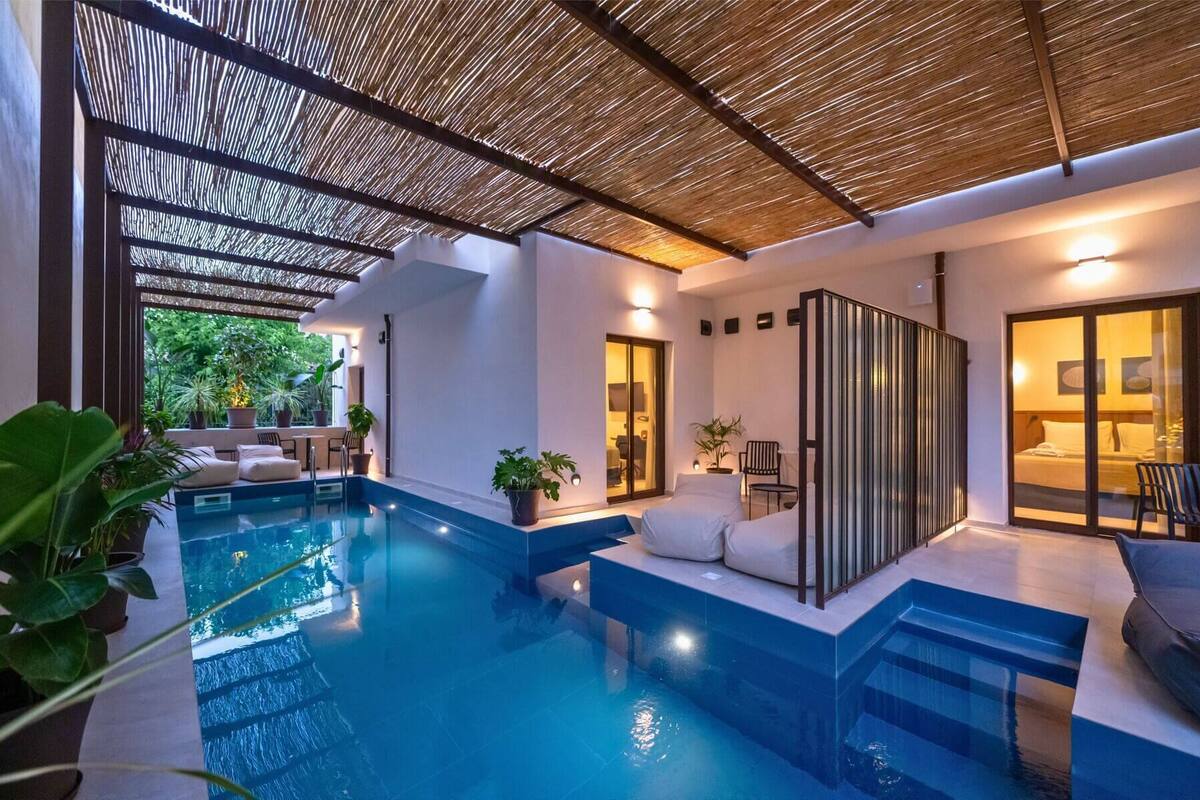Un área de piscina interior moderna con cerramiento de vidrio, zona de estar con sofá y sillones, y vistas al exterior.