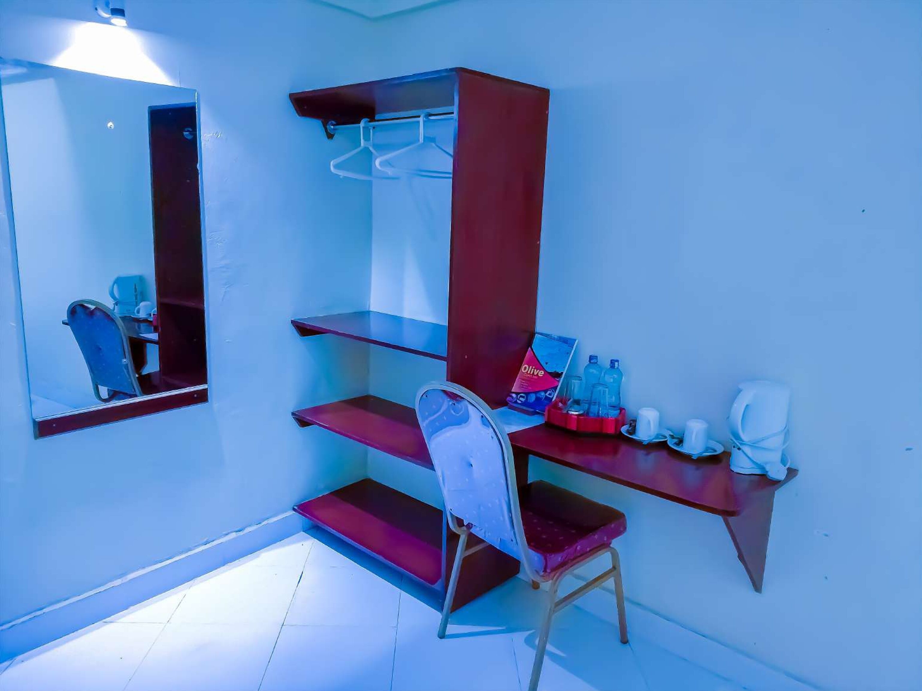 Una habitación pequeña y funcional con escritorio, silla y espejo. Hay estantes con algunos objetos, incluyendo una botella y una taza.