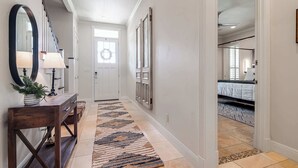 Un couloir avec une console en bois, un miroir et un tapis à motifs, qui mène à une chambre à coucher avec un lit et une fenêtre.