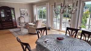 A dining area with a patterned tablecloth, a bowl of fruit, and a view of the outdoors through large windows.