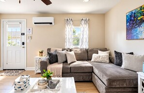 A living room with a sectional sofa, a coffee table with decorative items, a white door, and an air conditioning unit on the wall.