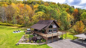 A two-story wooden cabin with a deck, surrounded by a well-maintained lawn and a forested hillside.