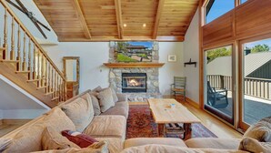A cozy living room with a stone fireplace, wooden ceiling, and a large sliding glass door to the outdoors.