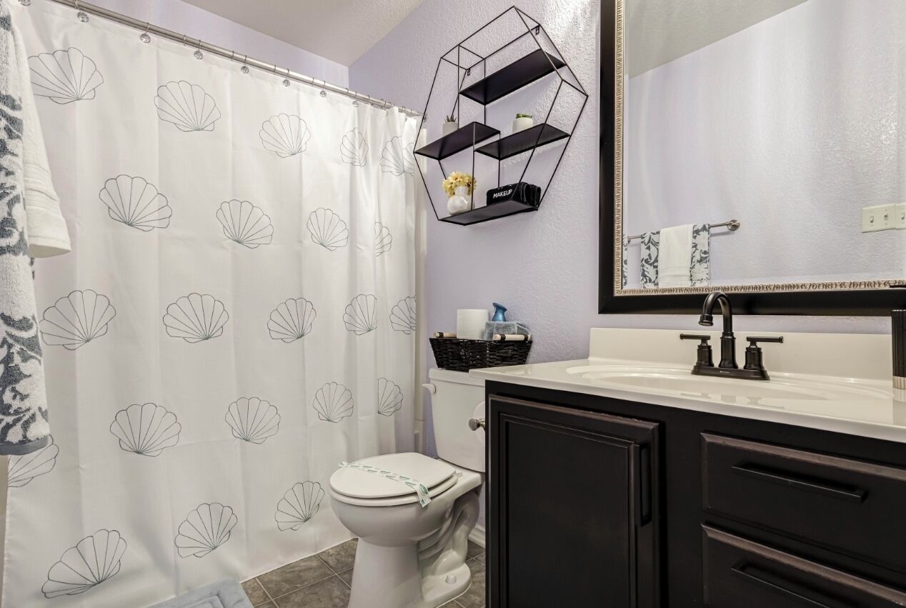 Une salle de bain comprenant des toilettes, un lavabo blanc avec un robinet noir, un miroir au-dessus du lavabo et un rideau de douche orné d’un motif de coquillages.