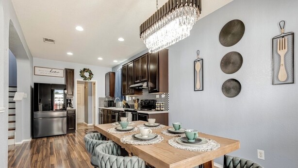A modern kitchen with dark wooden cabinets, a stainless steel refrigerator, and a dining area with a wooden table and grey upholstered chairs.