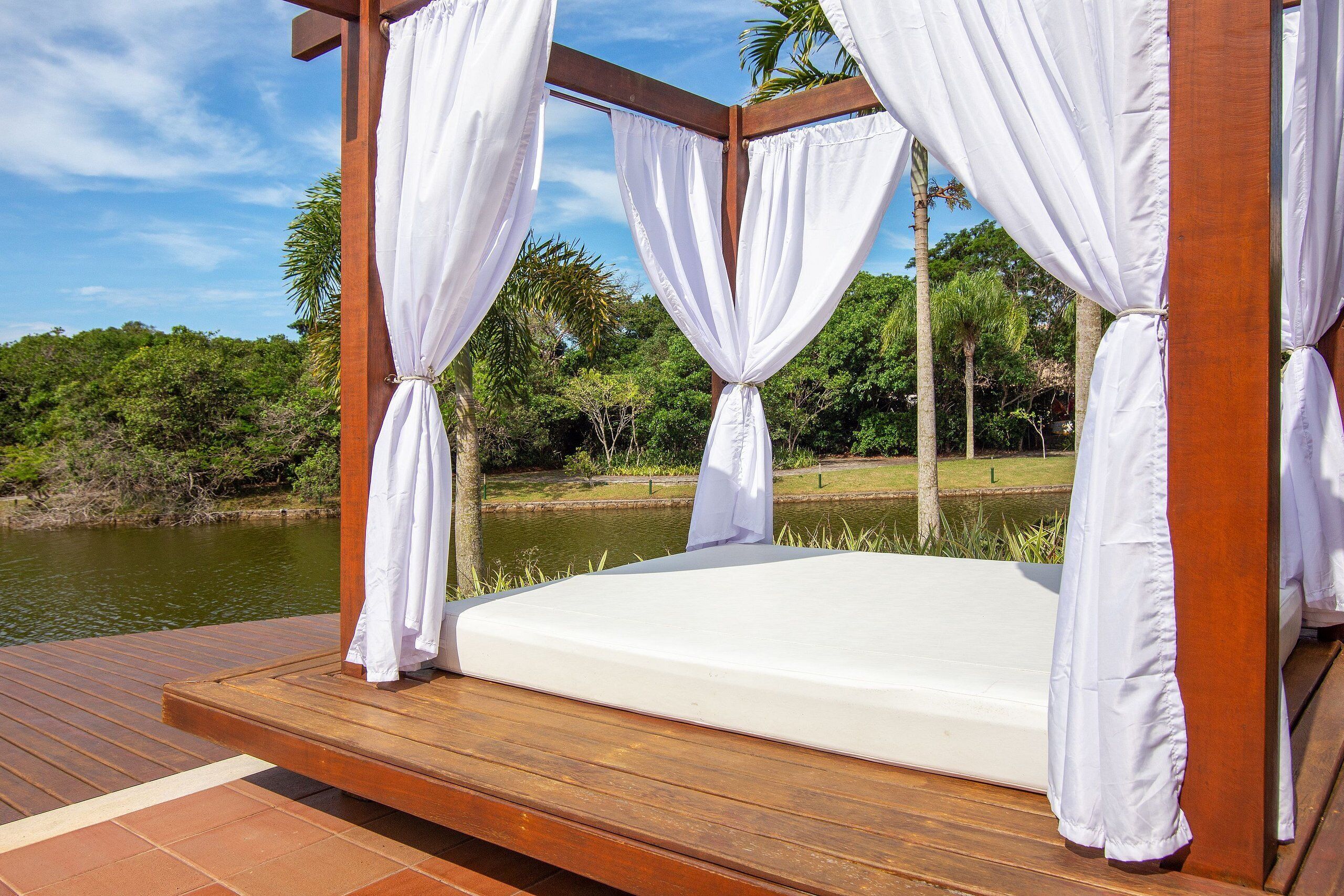 A wooden deck with a white canopy bed overlooking a body of water and lush greenery.