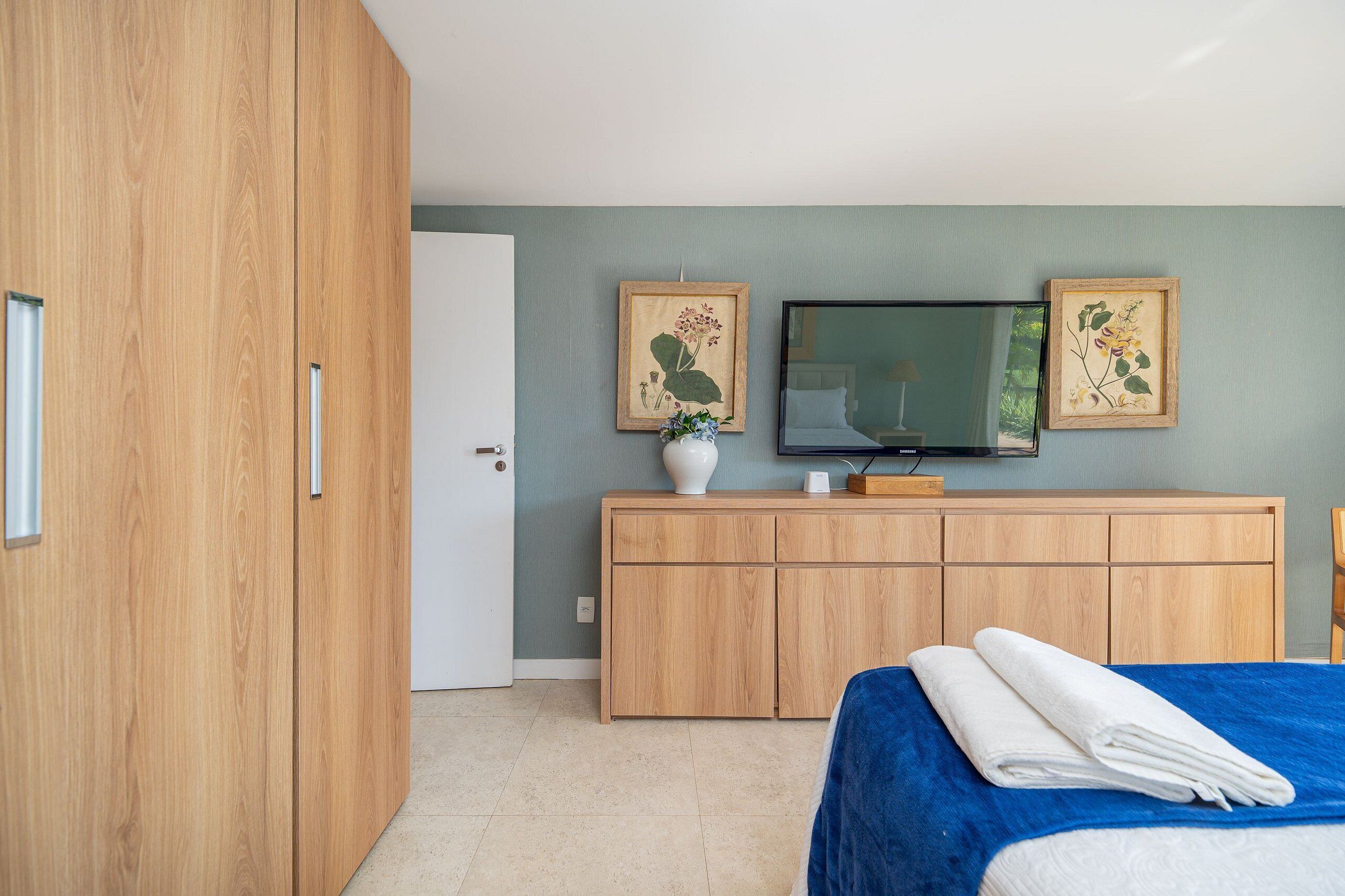 A bedroom with a bed, wooden wardrobe, a television, and two framed botanical prints on the wall.