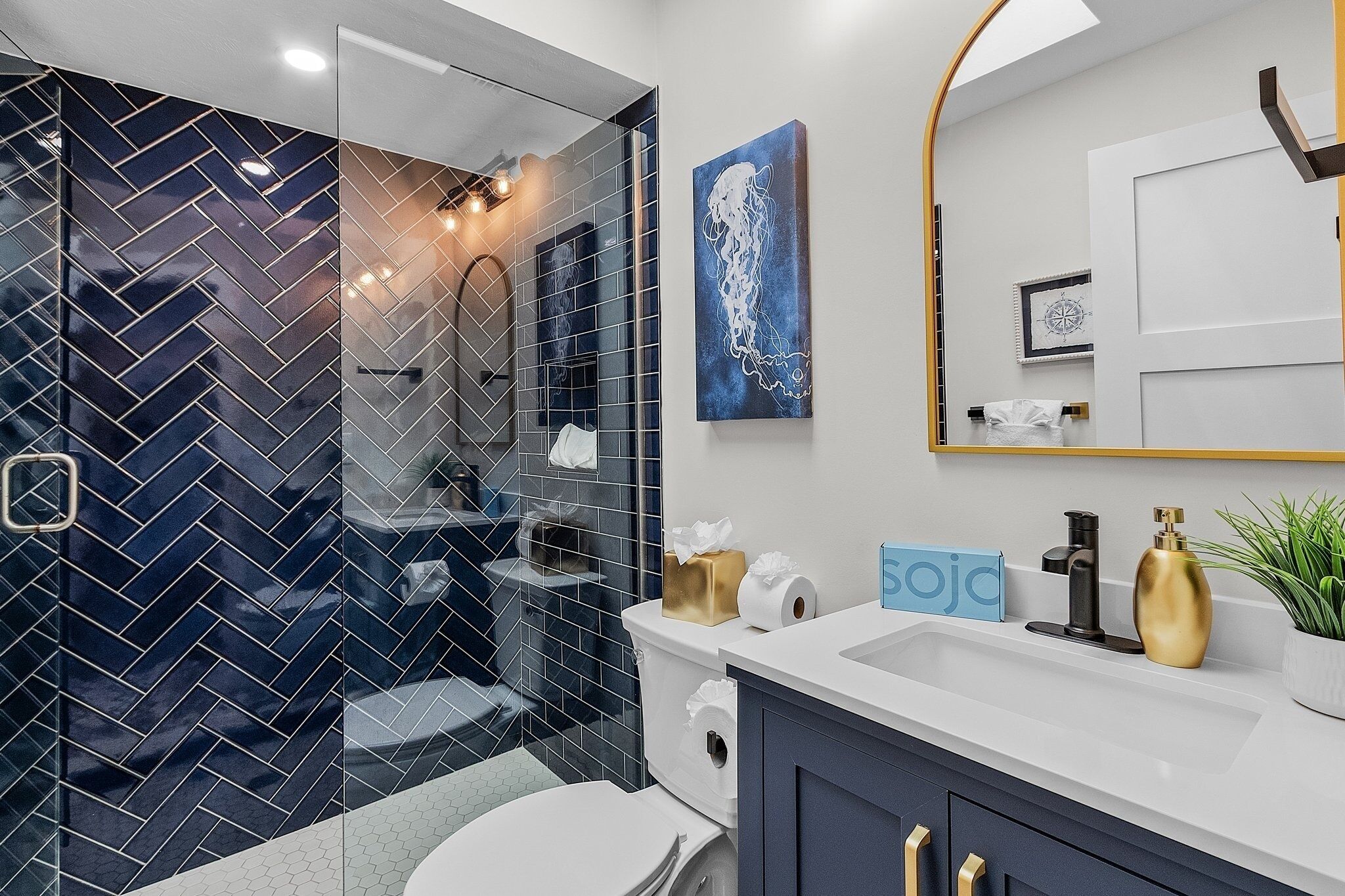 Ein modernes Badezimmer mit einer Duschkabine aus Glas, einer weißen Toilette, einem Waschbecken mit goldfarbenen Armaturen und einem blauen Kunstwerk an der Wand.