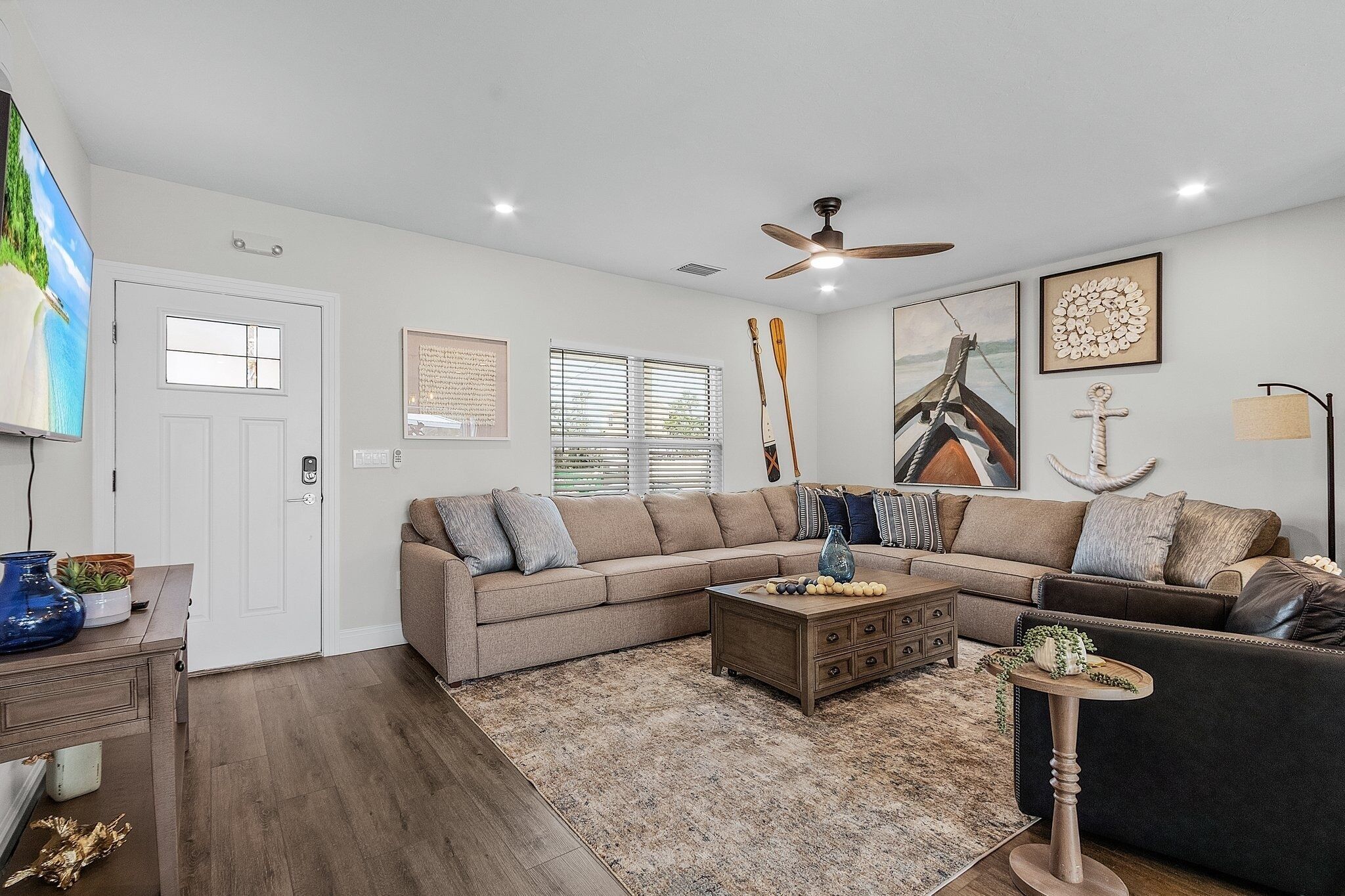 A living room with a sectional sofa, a coffee table, and a TV. There are windows with blinds, a ceiling fan, and wall art.