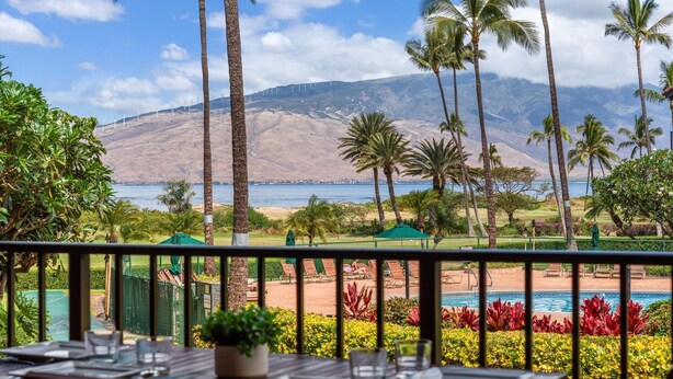 A dining area with a view of a beach, palm trees, and a mountain in the background. - Luana Kai C203 1 Bedroom Condo (Kihei)