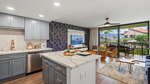 A modern kitchen with grey cabinets, a stainless steel dishwasher, and a large island. There is a balcony with outdoor furniture and a view of palm trees. - Luana Kai C203 1 Bedroom Condo (Kihei)
